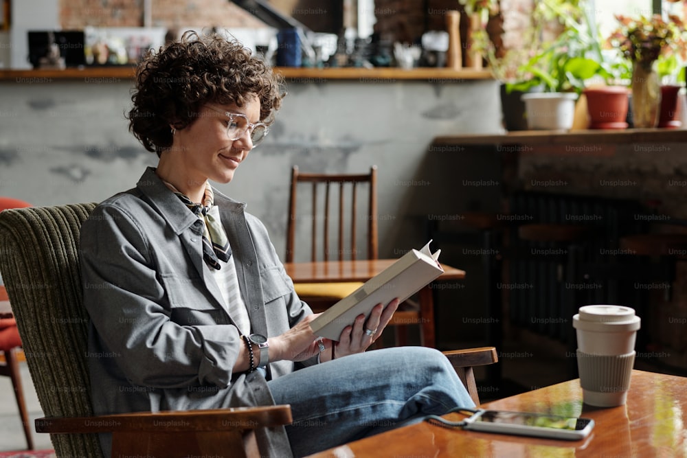 Glückliche junge Frau in Freizeitkleidung und Brille entspannt sich im Sessel mit interessantem Buch, während sie Freizeit im gemütlichen Café verbringt