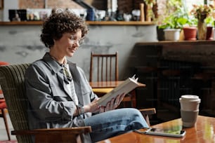 Mujer joven feliz en ropa casual y anteojos relajándose en un sillón con un libro interesante mientras pasa el ocio en un café acogedor