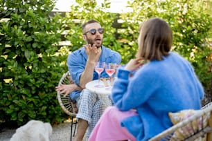 Young stylish couple have conversation while sitting by the round coffee table at their beautiful garden. Young man and woman have romantic evening outdoors