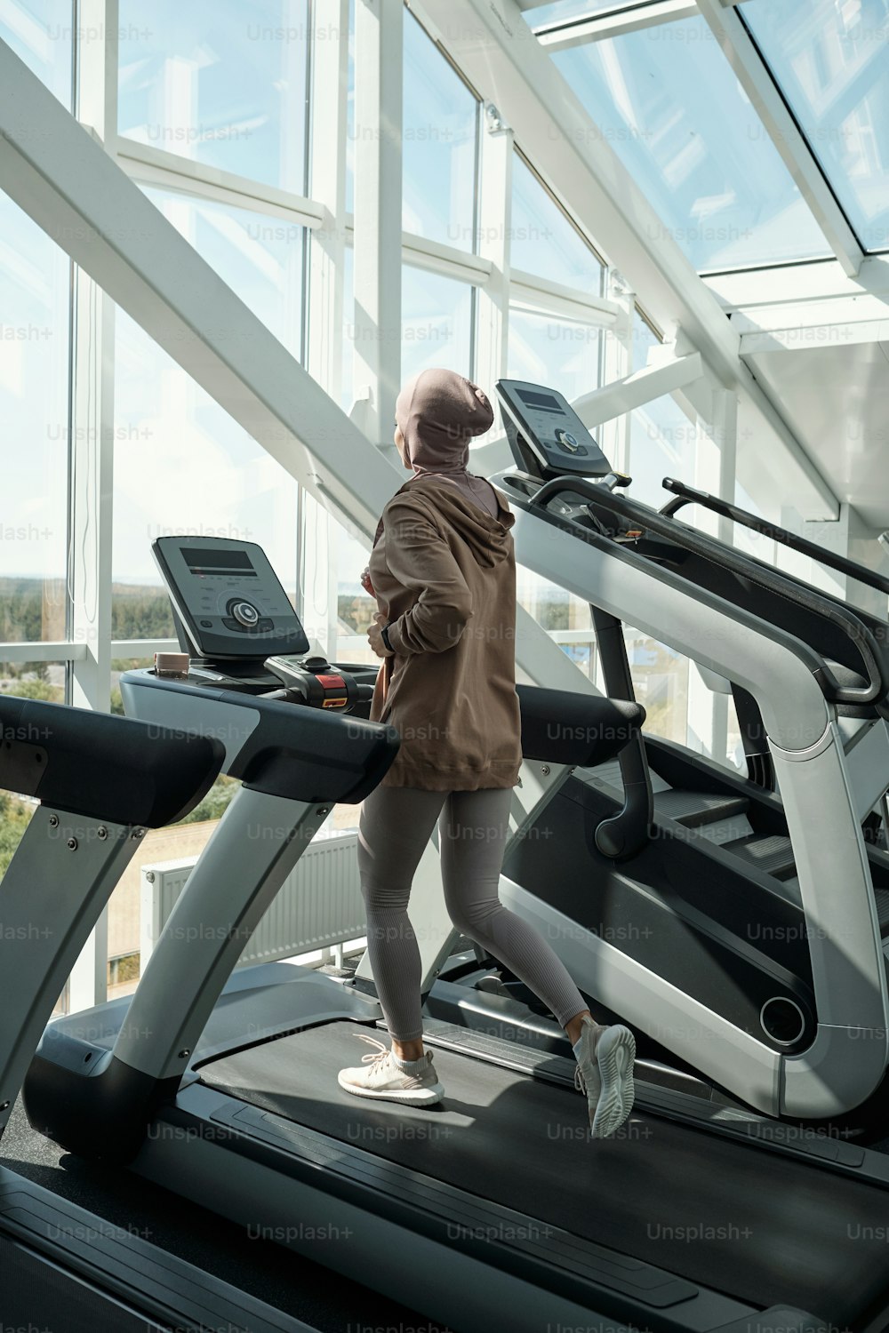 Vertikale lange Aufnahme einer nicht erkennbaren jungen Frau mit Hijab, die auf einem Laufband-Trainingsgerät im modernen Fitnessstudio läuft