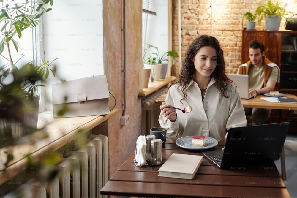 Giovane donna d'affari o libera professionista in abbigliamento casual che ha dessert mentre è seduta al tavolo davanti al computer portatile nel caffè e nel networking