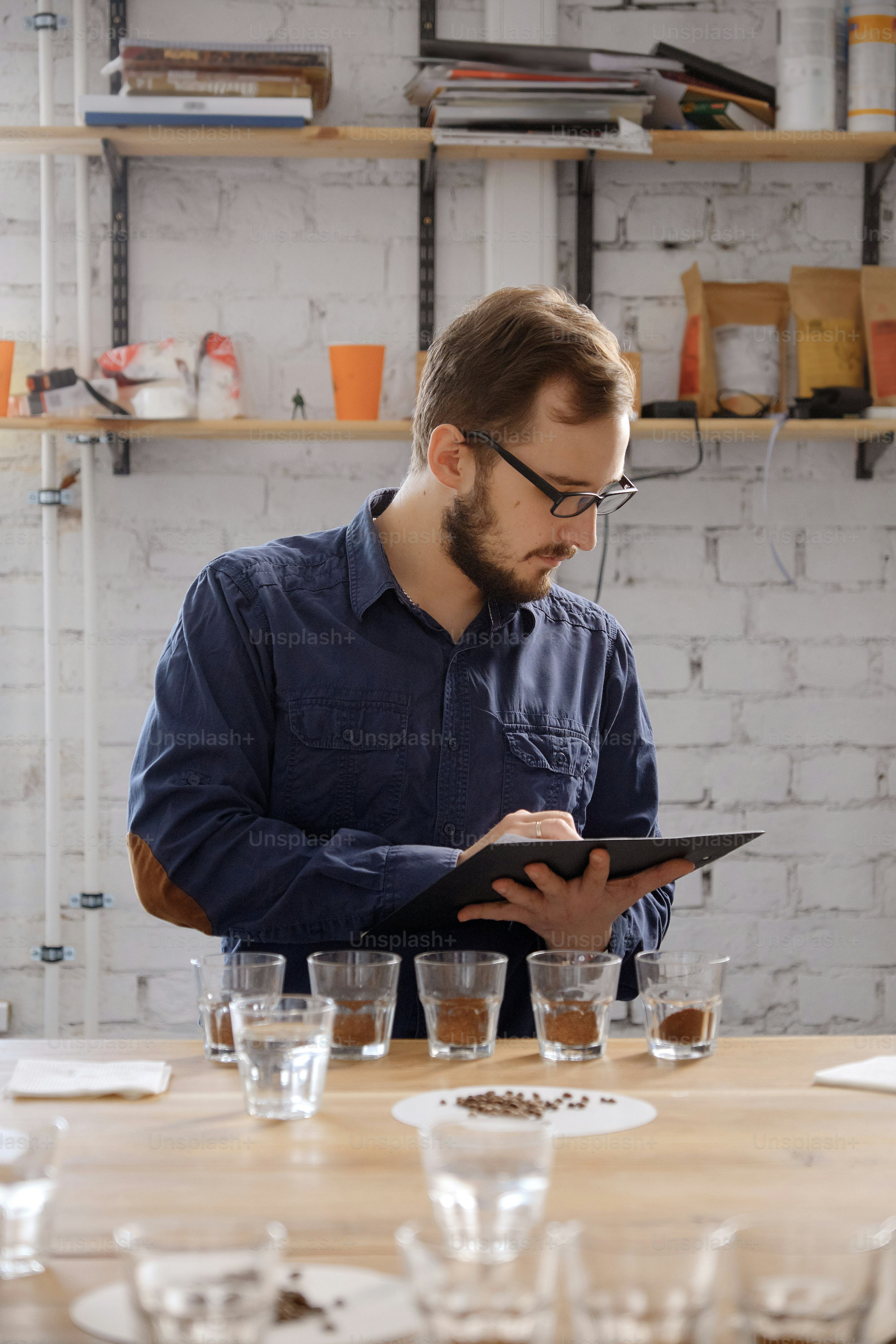 コーヒーカッピングテストの結果を書き、挽きたてのコーヒーの風味を調べる眼鏡をかけた男性の肖像画。彼は白い壁のそばに立っていて、ガラスのコップを持った列の前に立っている