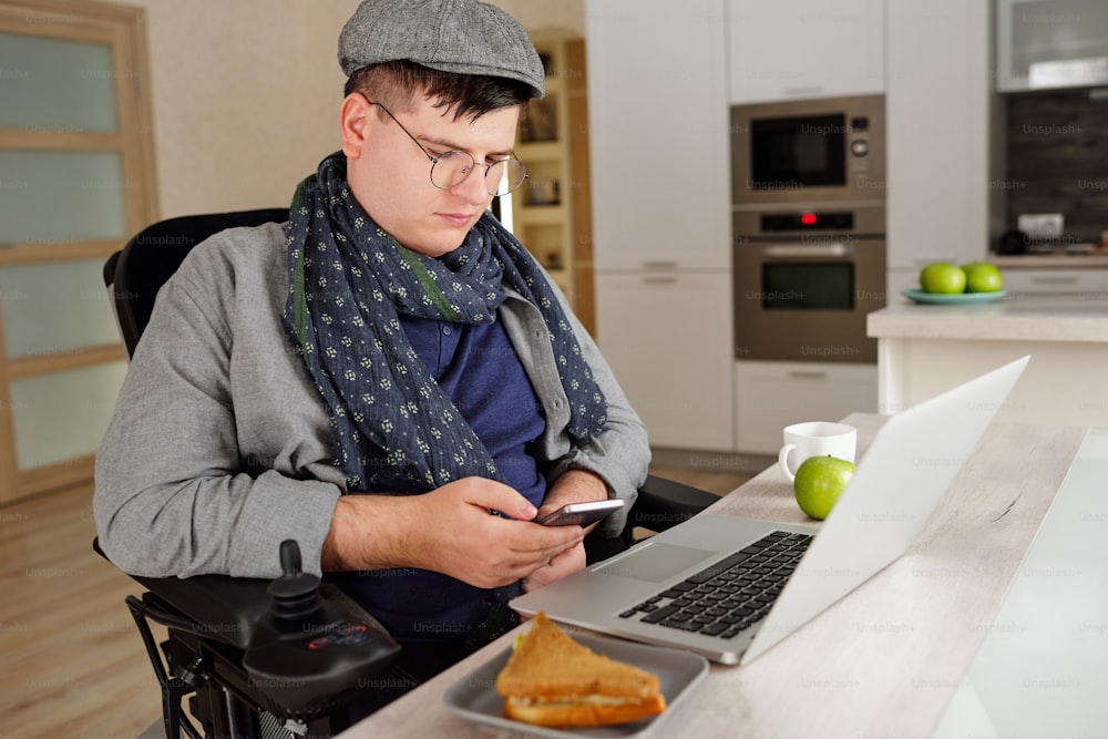 Young man in casualwear sitting in wheelchair while scrolling through contacts in smartphone in front of laptop