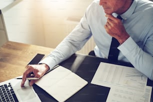 Planificador financiero maduro sentado en su escritorio en una oficina trabajando en línea con una computadora portátil y revisando documentos