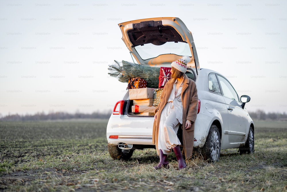 Donna in cappotto di pelliccia e cappello invernale seduta al bagagliaio dell'auto pieno di scatole regalo e albero di Natale sul campo al tramonto. Concetto di umore di Capodanno. Idea di preparazione per la vacanza