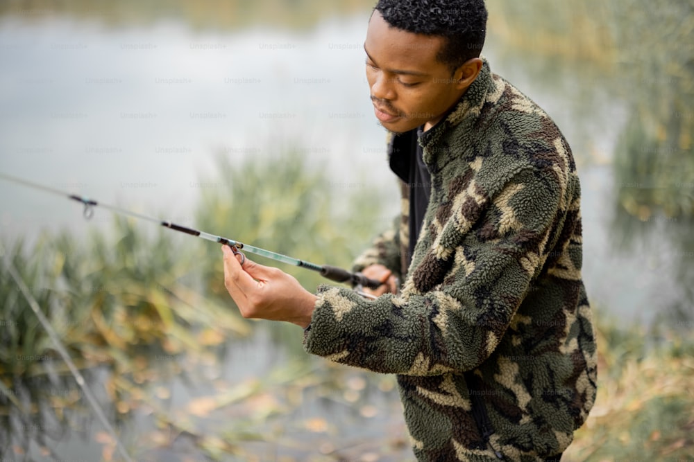 秋の季節に川や湖の海岸で釣りをしているときに釣り竿を修正するヒスパニック系の男性。自然の中での休息、趣味、週末のコンセプト。防寒着を着た男性の選択的焦点