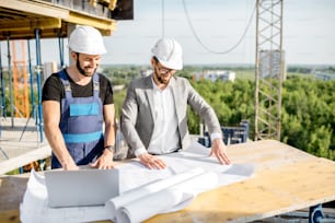 Ingénieur avec un travailleur en uniforme travaillant avec des dessins architecturaux et un ordinateur portable à la table sur le chantier de construction à l’extérieur