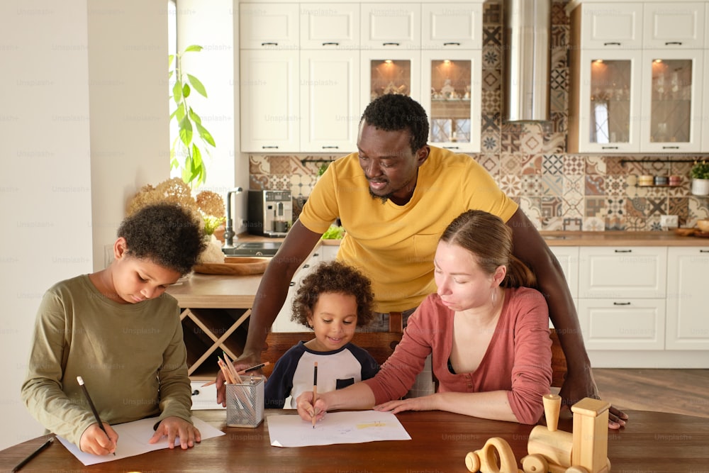 Giovane coppia interculturale e i loro due figli che disegnano con i pastelli dal tavolo di legno nell'ambiente della cucina