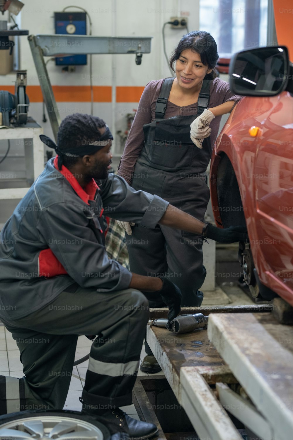 Vertical portrait of two ethnic mechanics repairing car in garage shop, focus on smiling young woman wearing workwear