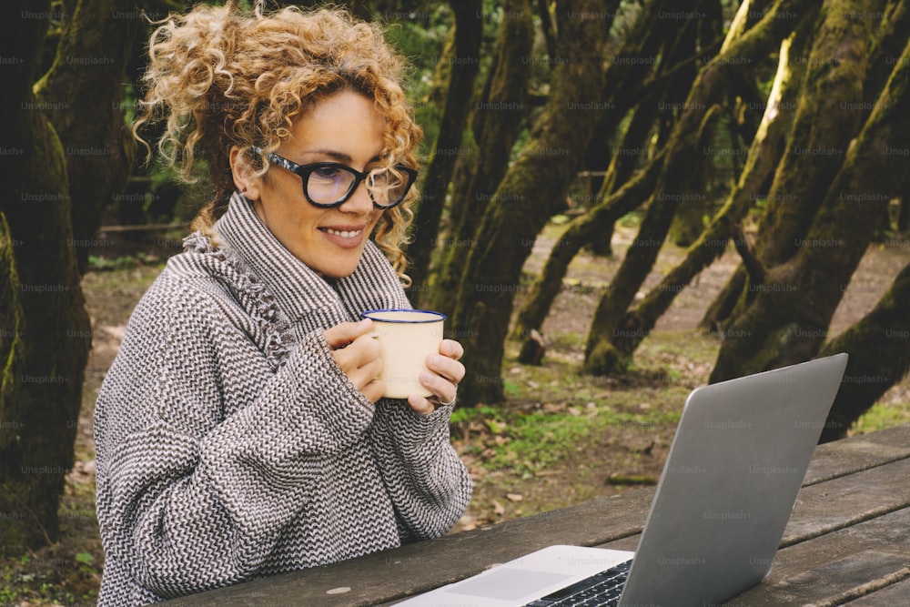 Attractive adult woman use laptop computer outdoor sitting in the trees forest outdoors. Concept of remote worker and digital nomad connection lifestyle. Female people use notebook and smile