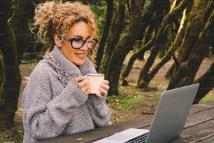 매력적인 성인 여성은 야외에서 나무 숲에 앉아 노트북 컴퓨터를 사용합니다. 원격 작업자와 디지털 유목민 연결 라이프 스타일의 개념. 여성들은 노트북을 사용하고 미소를 짓는다
