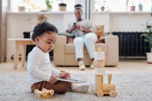 Pequeño hijo afroamericano tranquilo con camisa blanca sentado en el suelo y usando una tableta mientras juega solo con un juguete de madera