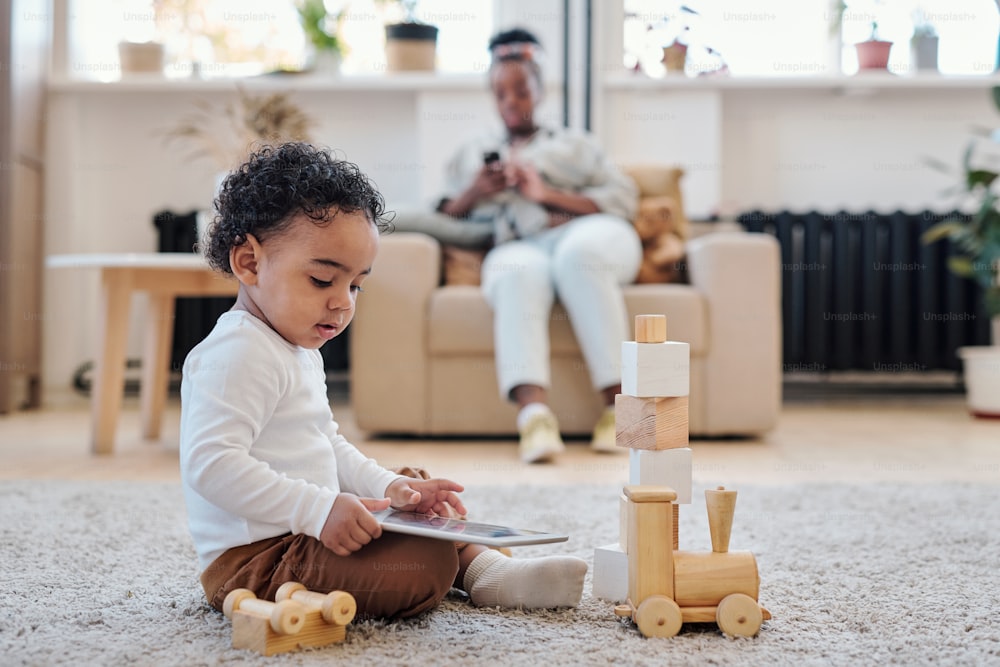 白いシャツを着た落ち着いたアフリカ系アメリカ人の息子が床に座り、一人で木のおもちゃで遊びながらタブレットを使用しています
