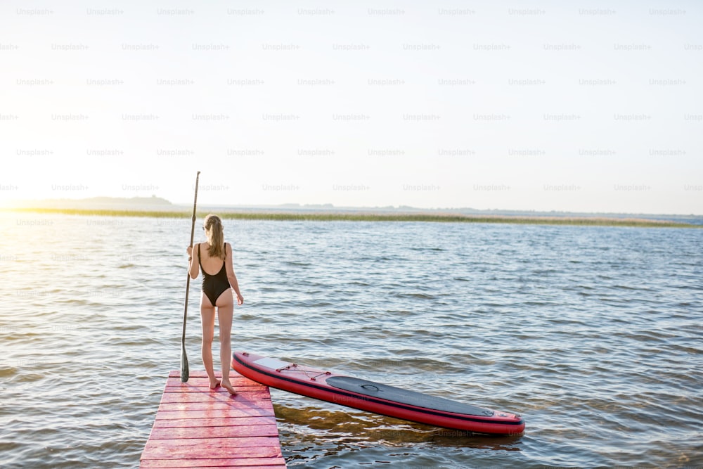 Beautiful woman standing back on the pier with paddleboard and oar enjoying morning view on the lake