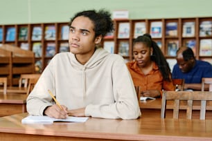 Joven negro pensativo sentado en el escritorio en el aula que tiene una lección en la escuela internacional para inmigrantes escuchando al maestro y tomando notas
