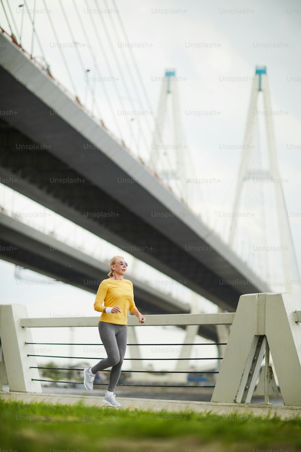 Fêmea idosa ativa vestindo pulôver amarelo e leggins cinzas correndo ao longo da ponte ao ar livre