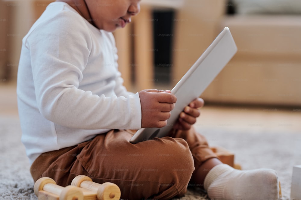Primer plano de un niño negro sentado con las piernas cruzadas en la alfombra y usando la aplicación de aprendizaje en la tableta