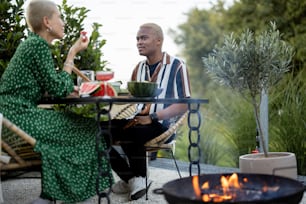 다인종 커플은 자연 위의 시골집 뒤뜰에서 저녁을 먹습니다. 건강한 식생활과 현대적��인 생활 방식에 대한 아이디어. 함께 시간을 즐기는 흑인 남성과 유럽 여성