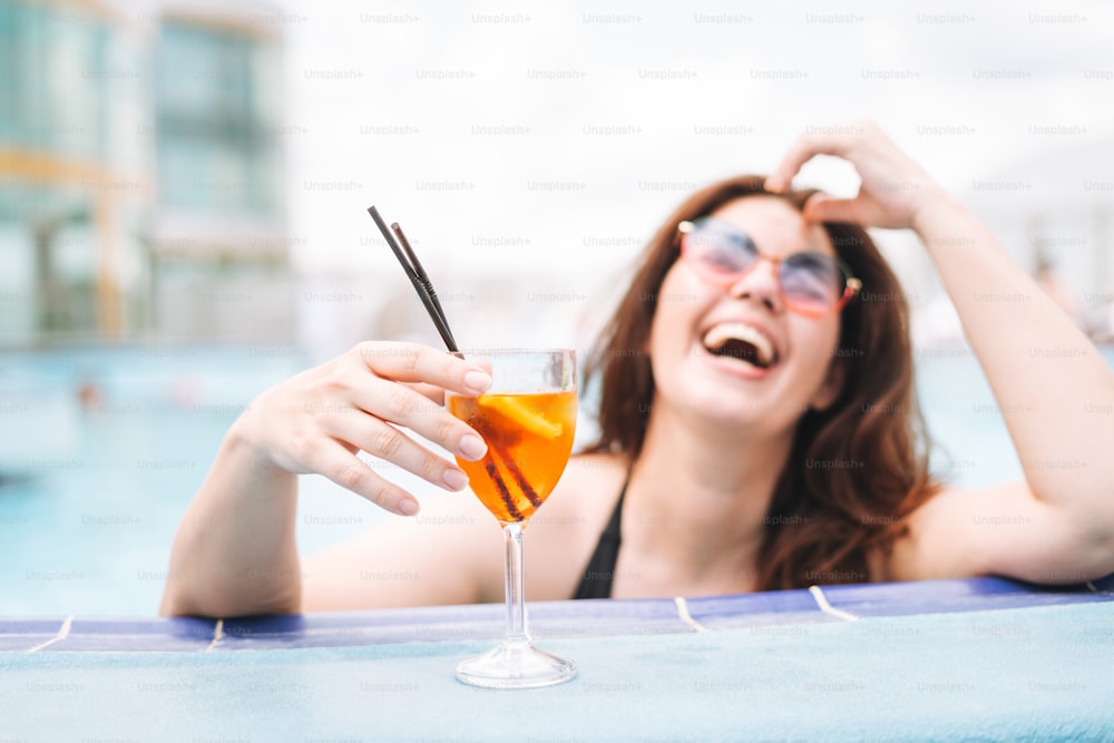Stylish happy laughing young woman plus size body positive in black swimsuit and sunglasses enjoying her life with glass of cocktail in hotel pool, selective focus