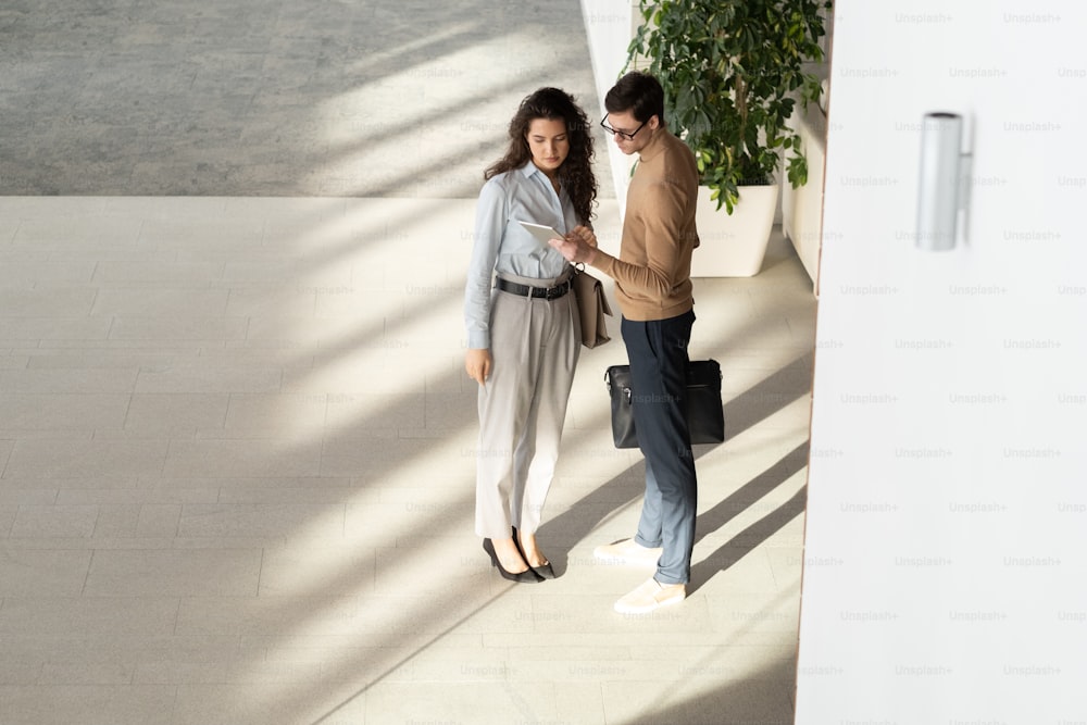 Young businessman with tablet making presentation of his analysis of data to female colleague at working meeting in large modern office