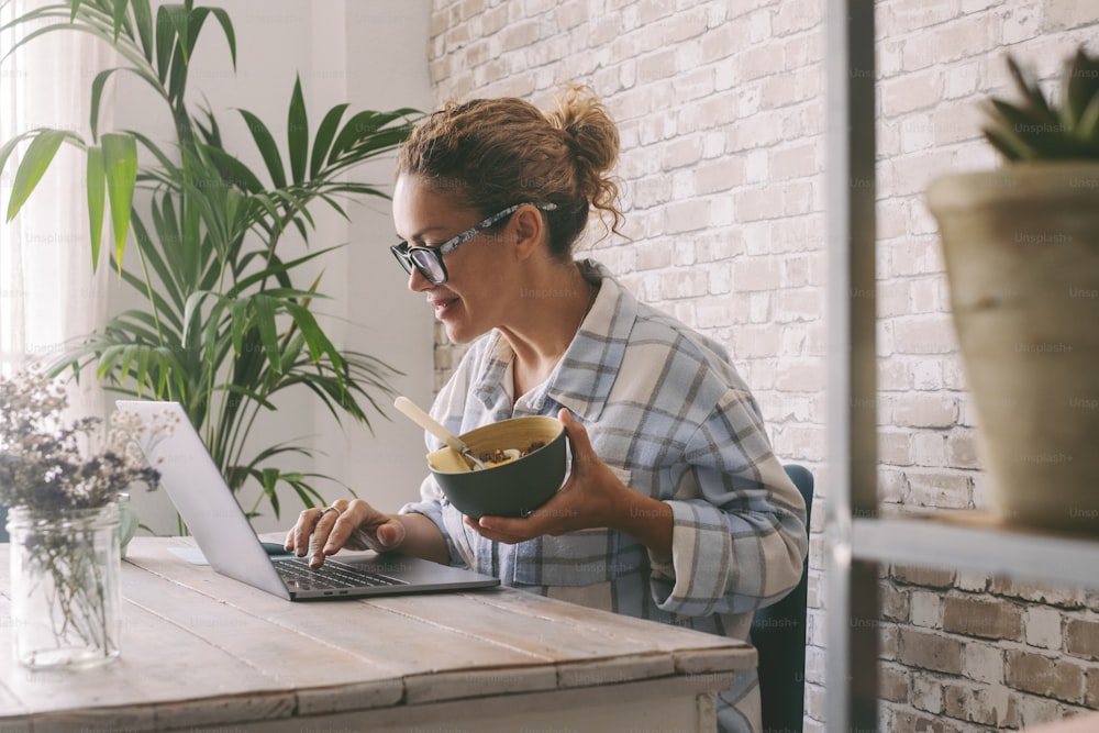 Femme travaillant à la table de bureau à domicile mangeant des aliments sains dans un bol. Mode de vie des femmes indépendantes, occupation alternative à l’aide d’un ordinateur portable et d’une opportunité en ligne. Entreprise ouvrière moderne