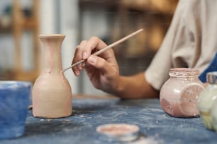 Close-up of unrecognizable potter using paintbrush while applying pink paint on clay vase in workshop