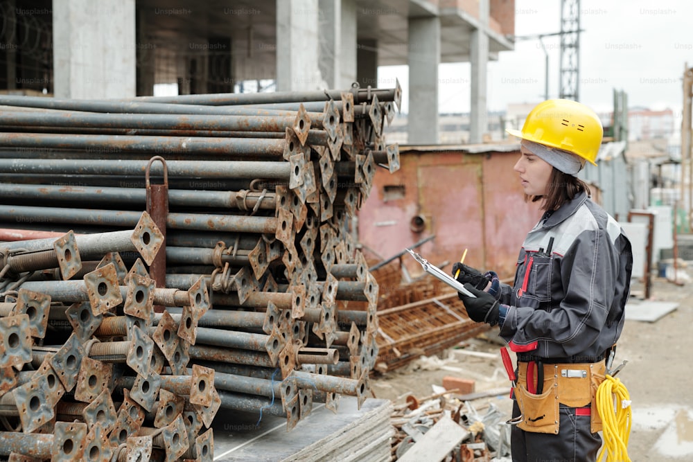 Young female engineer in workwear making notes in document while standing in front of two stacks of metallic pipes at construction area