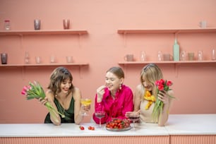 Beautiful caucasian girls celebrating women's day or having birthday party, standing together with alcohol drinks, fruits and flowers at kitchen bar indoors. Concept of spring time and holidays