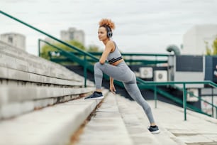 Una sportiva magra con i capelli ricci che ascolta musica in cuffia e si sgranchisce le gambe in piedi sulle scale in una zona urbana della città. Sportiva urbana che si allena all'aperto