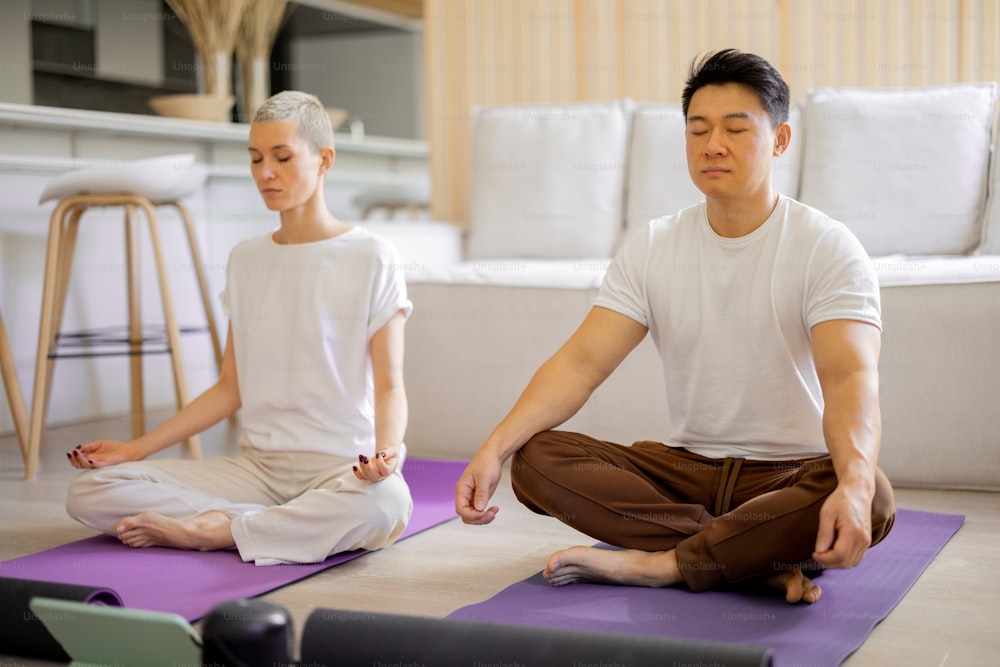 Multirassisches Paar, das Yoga praktiziert und meditiert, während es zu Hause auf Fitnessmatten sitzt. Konzept des gesunden Lebensstils. Idee von häuslichem Hobby und Freizeit. Asiatischer Mann und kaukasisches Mädchen mit geschlossenen Augen