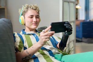 Junge lächelnde Frau in Freizeitkleidung schaut auf den Bildschirm des Smartphones in ihren Händen, während sie sich auf dem Sofa entspannt und im Video-Chat kommuniziert