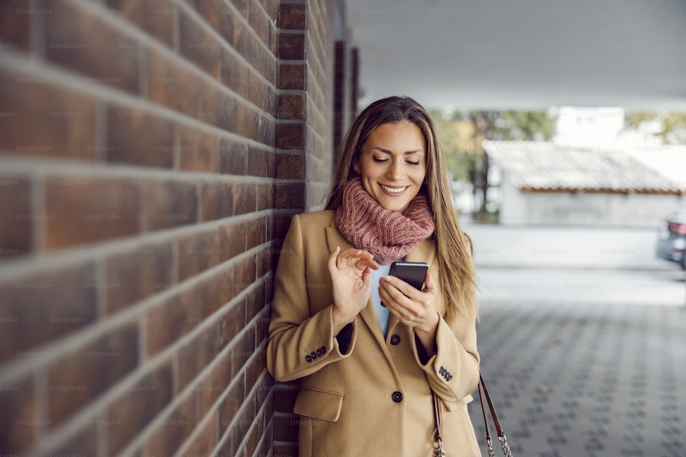현대의 온라인 데이트. 따뜻한 옷을 입은 젊은 행복한 여자가 벽돌 벽에 기대어 스마트폰을 사용하여 데이트 플랫폼에 액세스합니다.