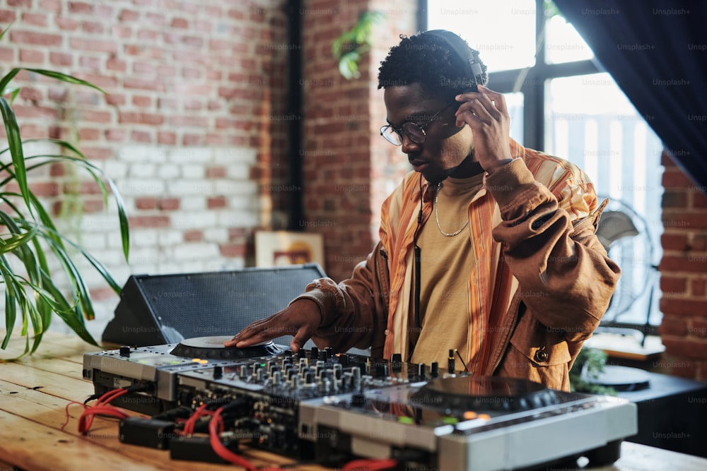 Black man in eyeglasses, casualwear and headphones making new music, listening to it and recording while standing in front of turntables