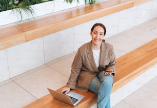 Erwachsene lächelnde brünette Geschäftsfrau vierzig Jahre mit langen Haaren in stilvollem beigefarbenem Anzug und Jeans arbeitet am Laptop am öffentlichen Platz