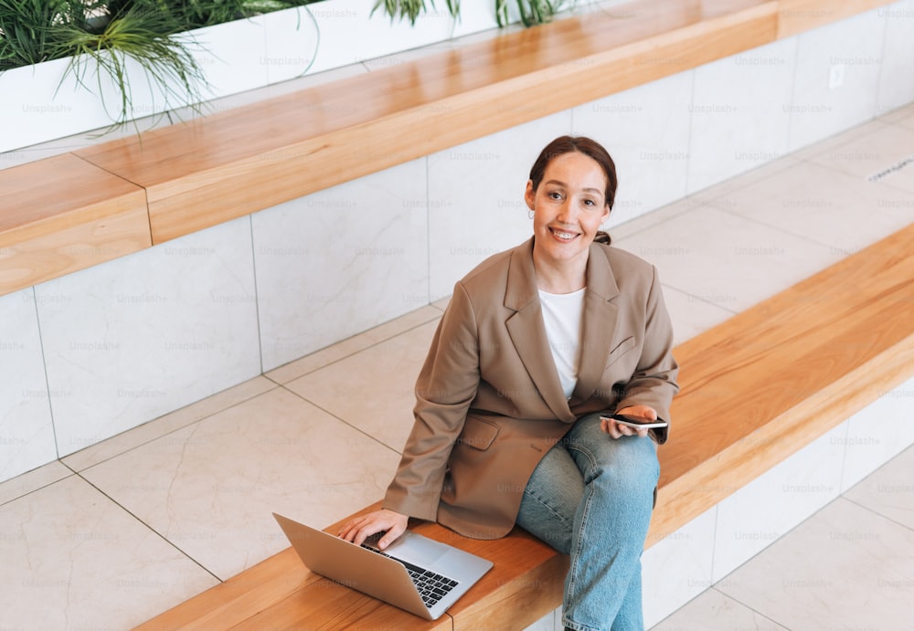 Mujer de negocios morena sonriente adulta de cuarenta años con cabello largo en elegante traje beige y jeans trabajando en computadora portátil en el lugar público