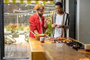 Two brightly dressed stylish guys having fun while making salad together on background of backyard. Concept of gay couples and everyday life at home. Caucasian and hispanic man cooking healthy food