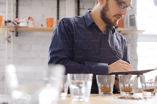 Retrato de un hombre con gafas escribiendo los resultados de la prueba de catación de café, examinando el café molido fresco para el sabor. Está parado cerca de una pared blanca frente a filas con vasos de vidrio