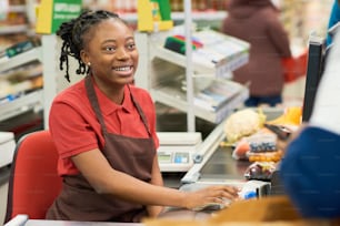 Happy young cashier in workwear looking at customer while sitting by workplace against display and counter line with food products