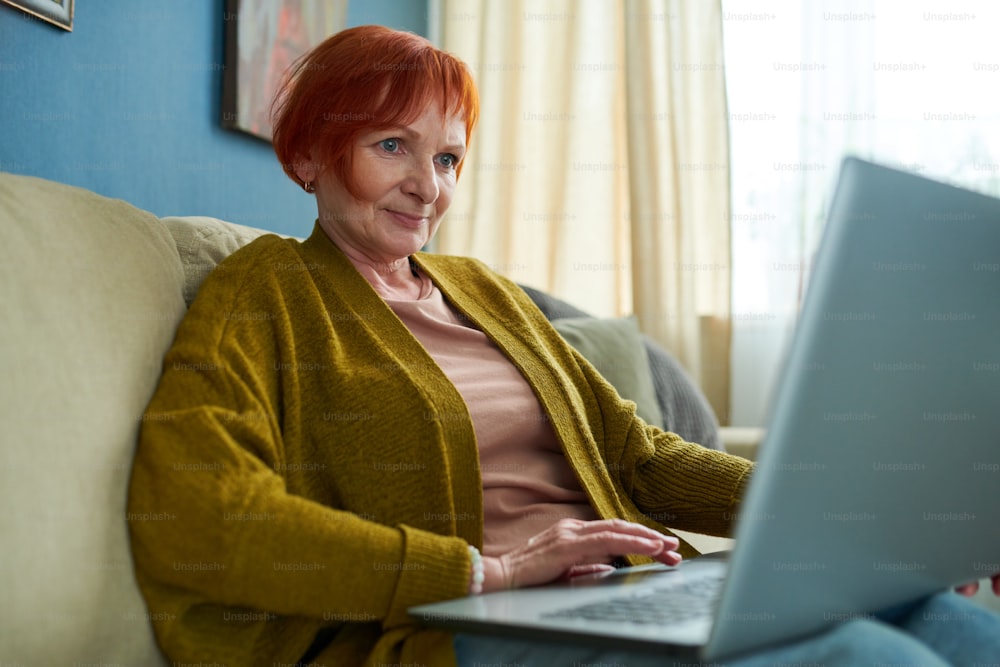 Retired woman with red hair sitting on sofa in living room and typing on laptop