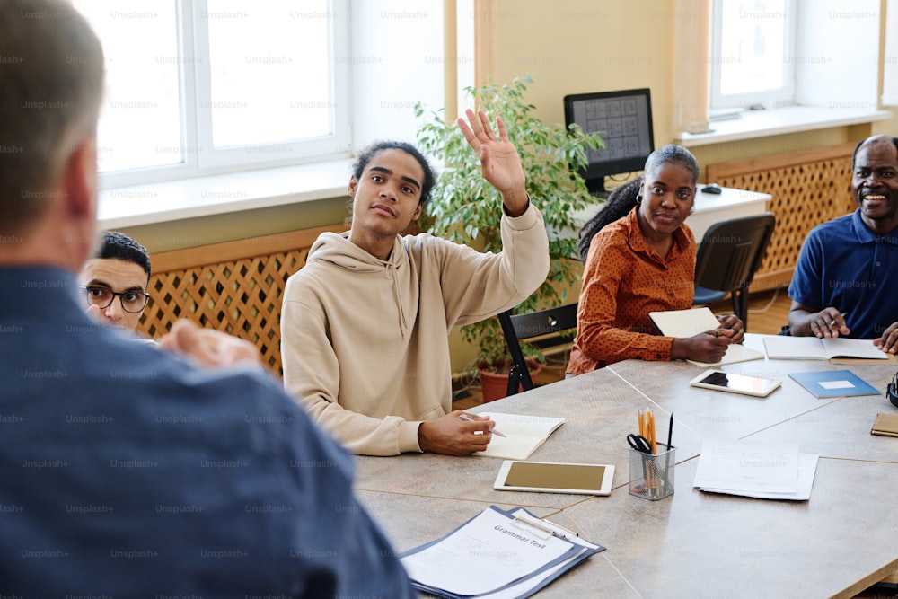 Joven estudiante negro inmigrante que asiste a clases de idiomas sentado en la mesa levantando hanf para hacer preguntas al maestro
