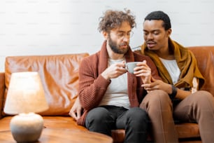 Gay couple with different nationality having close conversation while sitting with hot drinks on a couch at home. Concept of homosexual relations and lifestyle at home