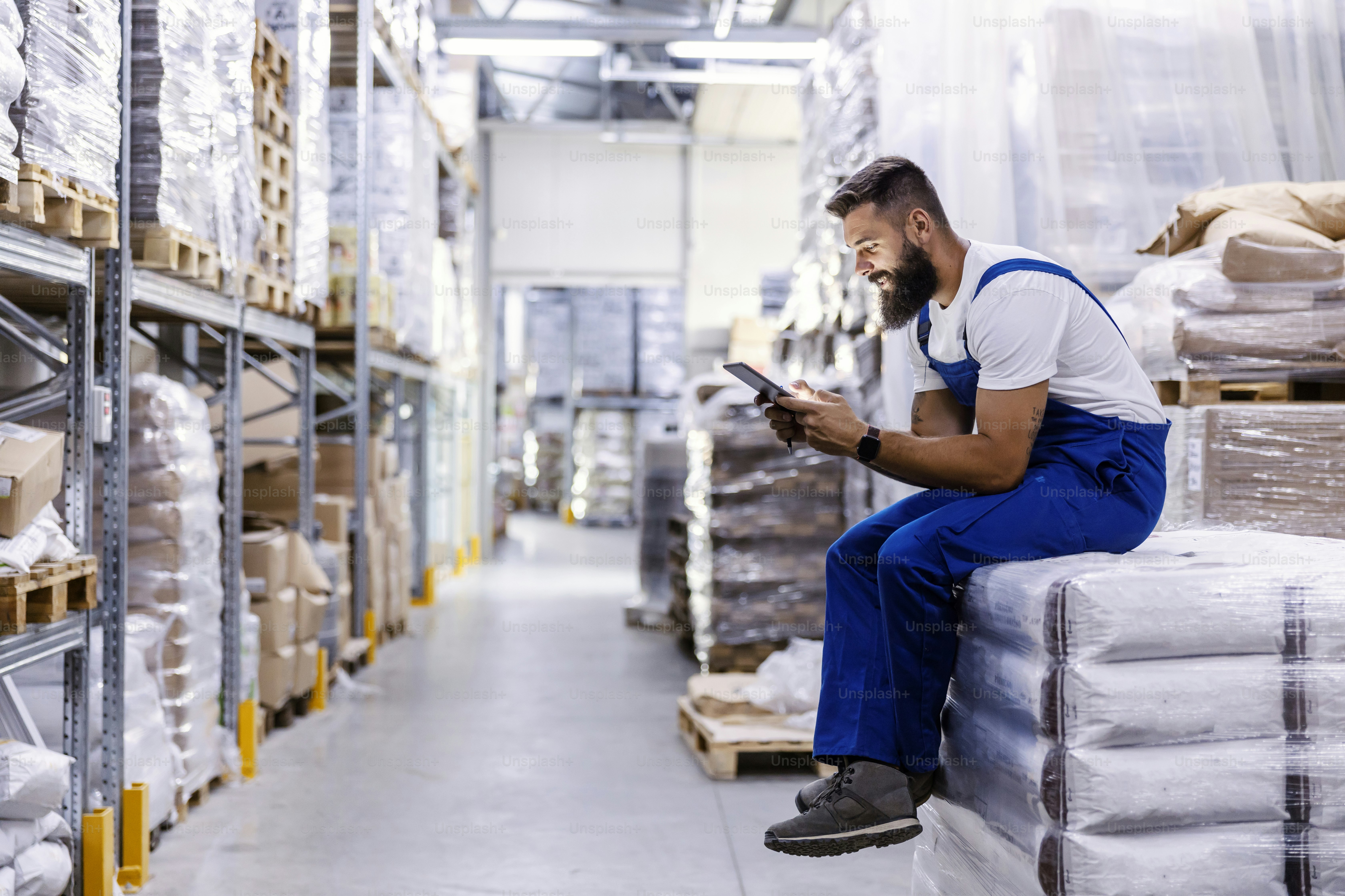 The storage worker is sitting on the pile of bags with flour and he is using the tablet for checking on orders. The orders for sipping can arrive to a client in just one day.