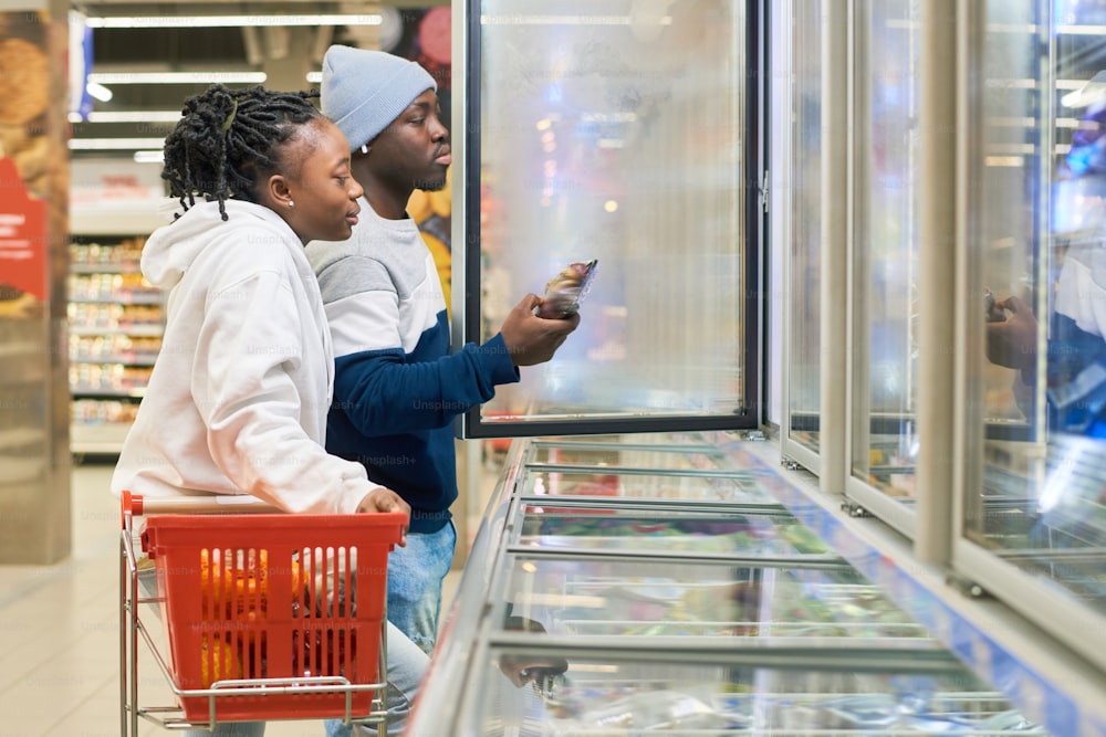 Jeune couple avec caddie choisissant des produits alimentaires surgelés dans un supermarché pendant qu’un gars sort un paquet de fruits du réfrigérateur