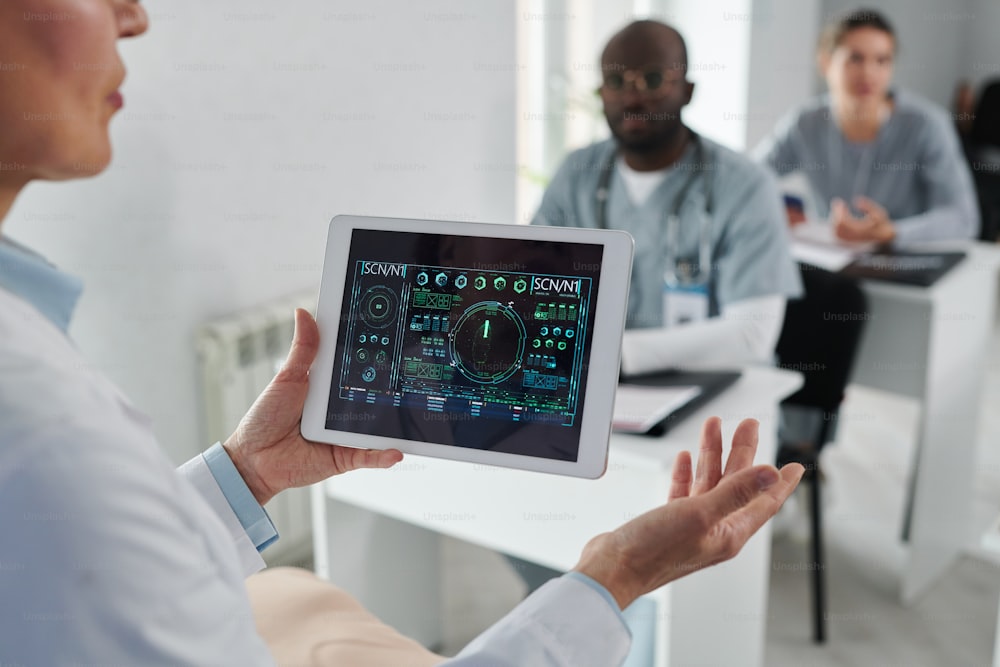 Close-up de professor maduro segurando tablet digital com gráficos em suas mãos e conversando com os alunos durante o treinamento médico