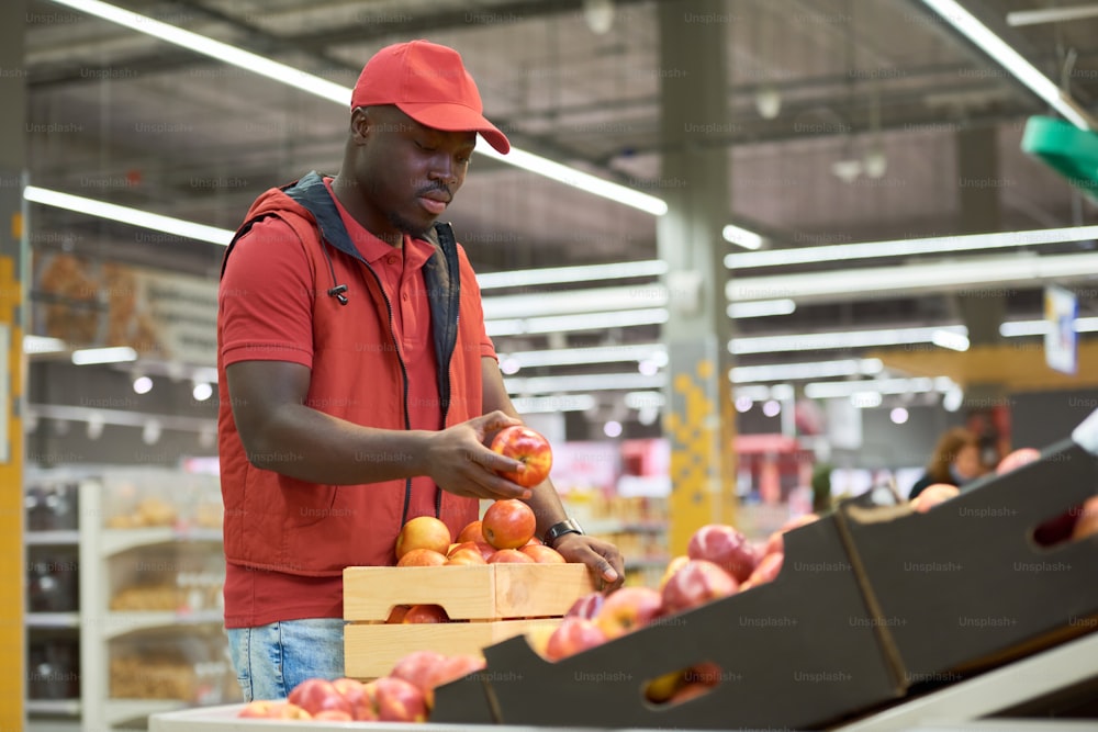 Black man in red uniform puting fresh ripe apples from wooden box on display with other fruits and vegetables in grocery department