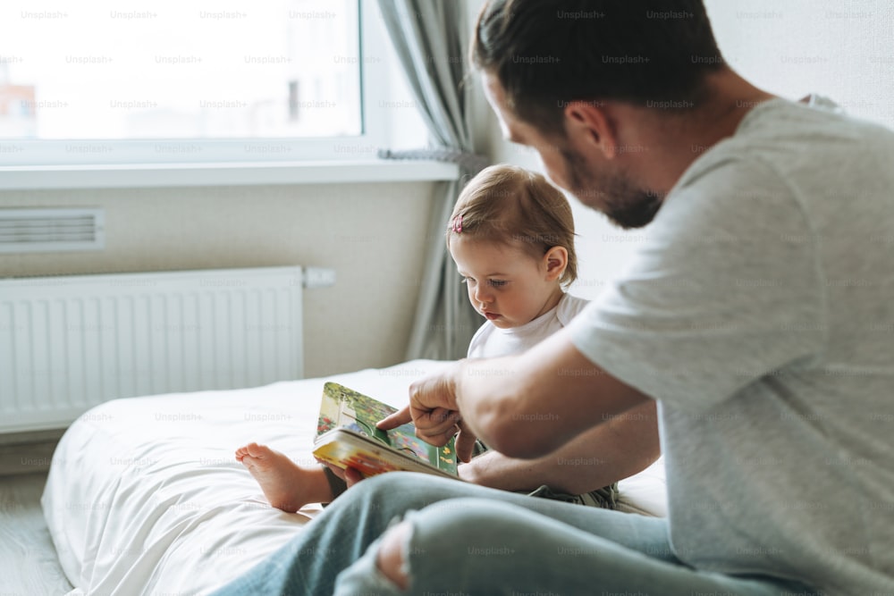 Happy father young man and baby girl little daughter having fun reading a book in children room at home