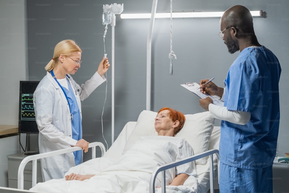 El médico prescribe medicamentos mientras la enfermera hace un gotero, tratan a una mujer mayor en la sala del hospital