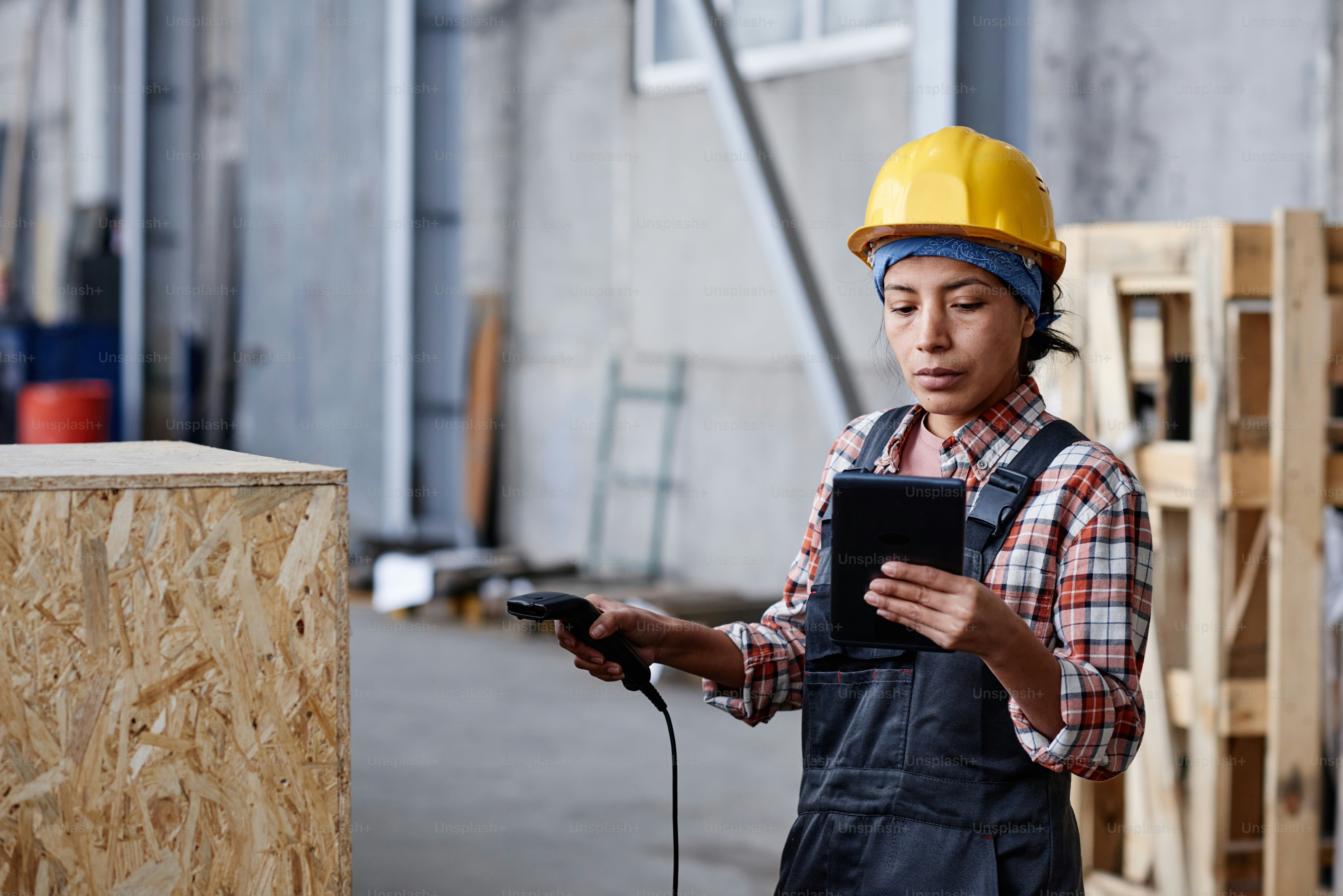 Young serious woman in coveralls and hardhat scanning barcode on huge box and looking at data on screen of tablet