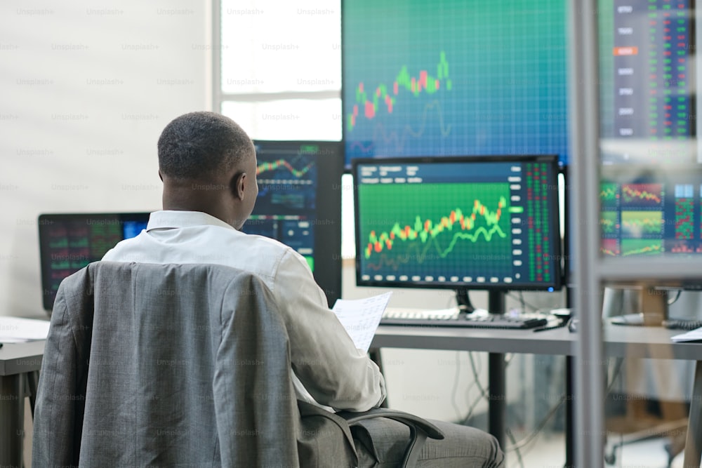 Vista trasera de un hombre afroamericano irreconocible sentado frente a monitores de computadora que se agitan con estadísticas monetarias