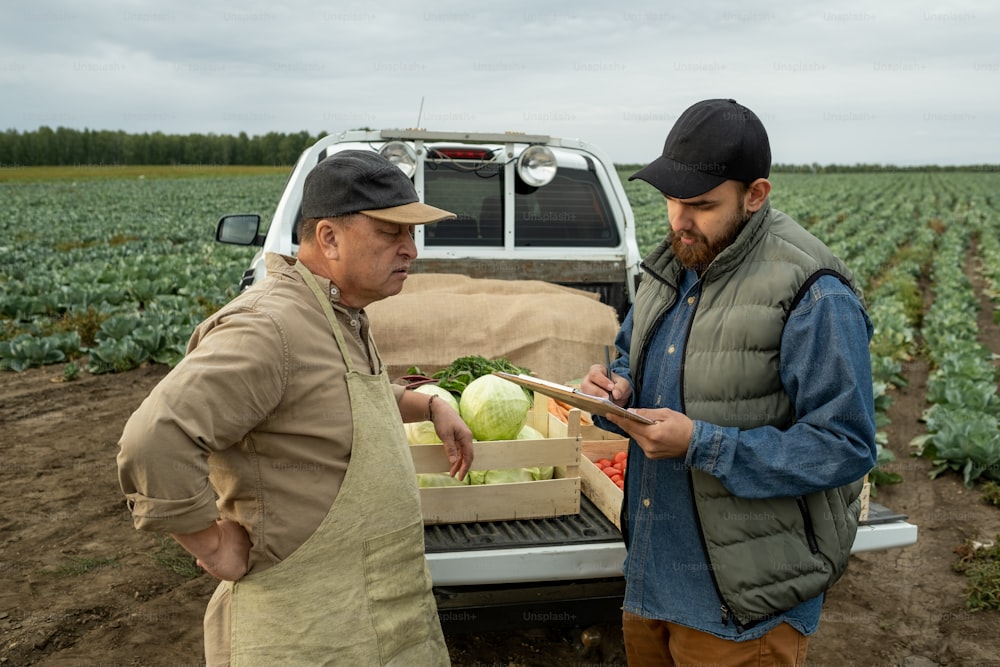 Mature farmer talking to young bearded man making notes in document